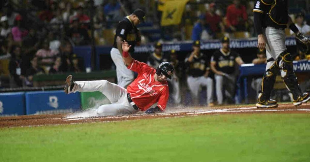 Yariel Rodríguez, a baseball player in a red uniform, slides into home plate, causing dirt and water to splash around him as players in black uniforms observe.